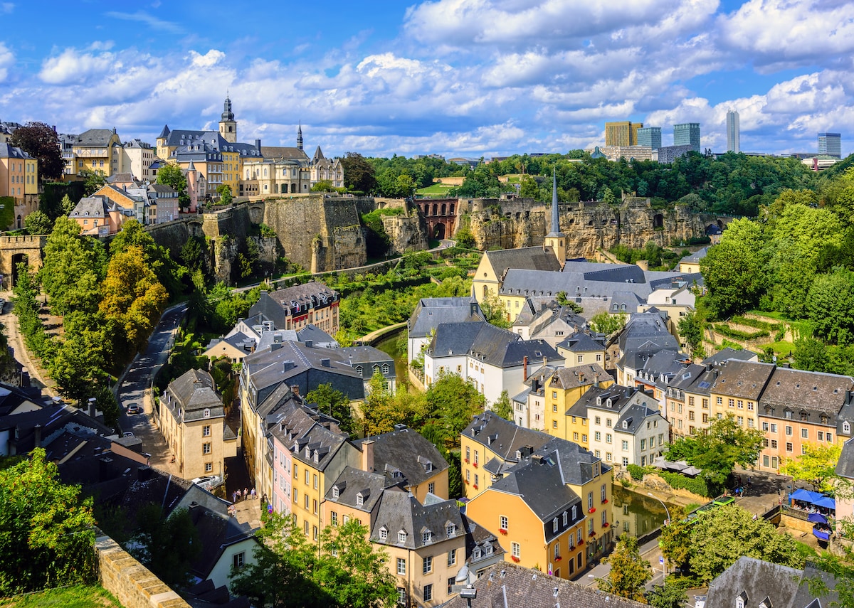 A panoramic view showcases a blend of historic and modern architecture in the city. Colorful buildings with sloped roofs are nestled among lush greenery, while the skyline features a mix of traditional structures and contemporary towers, framed by a vibrant blue sky with scattered clouds.