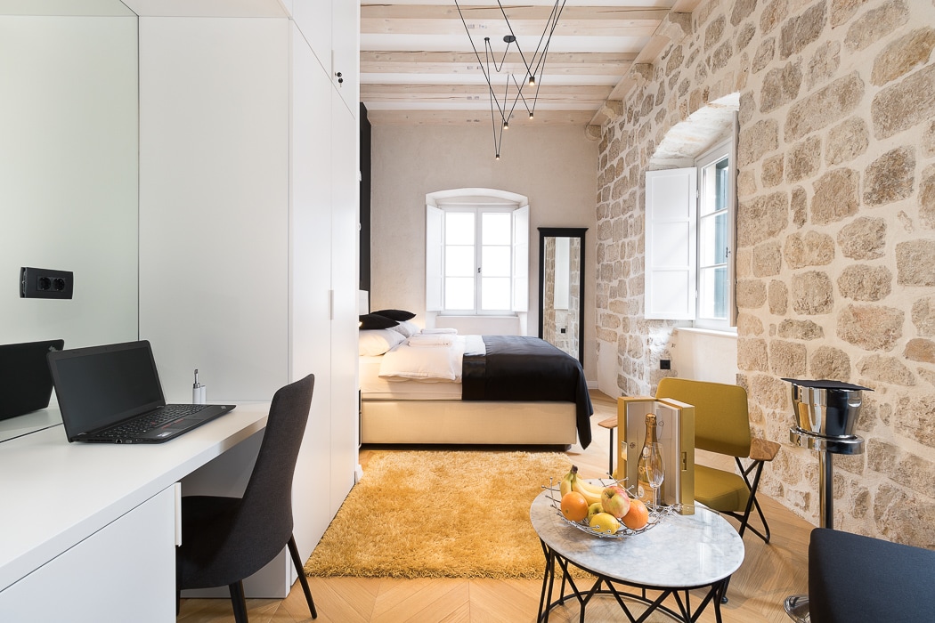 A serene room features a plush double bed with premium linens, set against a backdrop of exposed stone walls. A sleek work desk is positioned nearby, while a cozy seating area includes two armchairs and a small round table topped with fruit. Natural light enters through two windows.