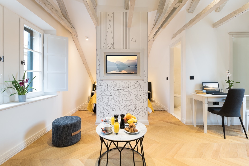 A bright studio space is highlighted by vaulted ceilings and natural light from the window. A small round coffee table is elegantly set with breakfast items, while a sleek desk and chair are positioned nearby. Decorative elements add character to the walls.