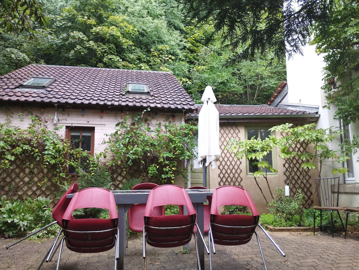 A tranquil outdoor space is presented, featuring a rectangular table surrounded by four red chairs. The backdrop includes a small wooden house and a neighboring structure, both partially covered by greenery. A large umbrella provides shade, enhancing the inviting atmosphere.