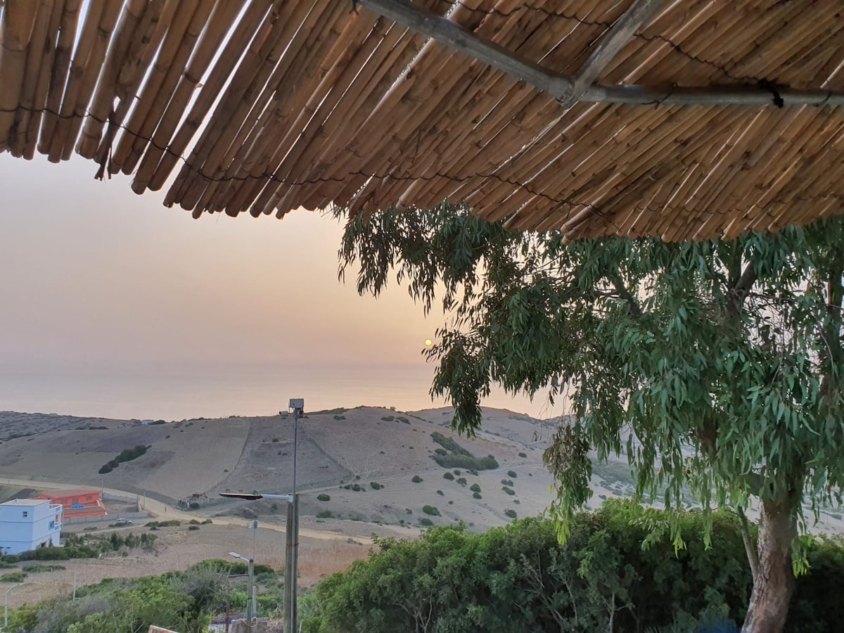 A shaded outdoor area is framed by a bamboo roof, offering views of the rolling hills and distant sea. The soft hues of a sunset are visible over the landscape, creating a calming atmosphere. Nearby foliage adds a natural touch to the scene.