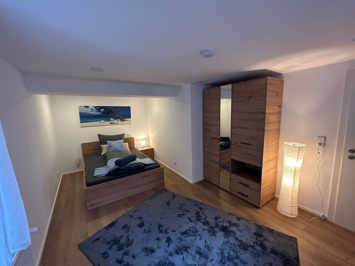 The bedroom features a modern wooden bed with neutral bedding, complemented by a decorative picture of a boat on the wall. A tall wardrobe provides storage, while a soft rug adds comfort underfoot. Soft lighting from a floor lamp enhances the peaceful ambiance.