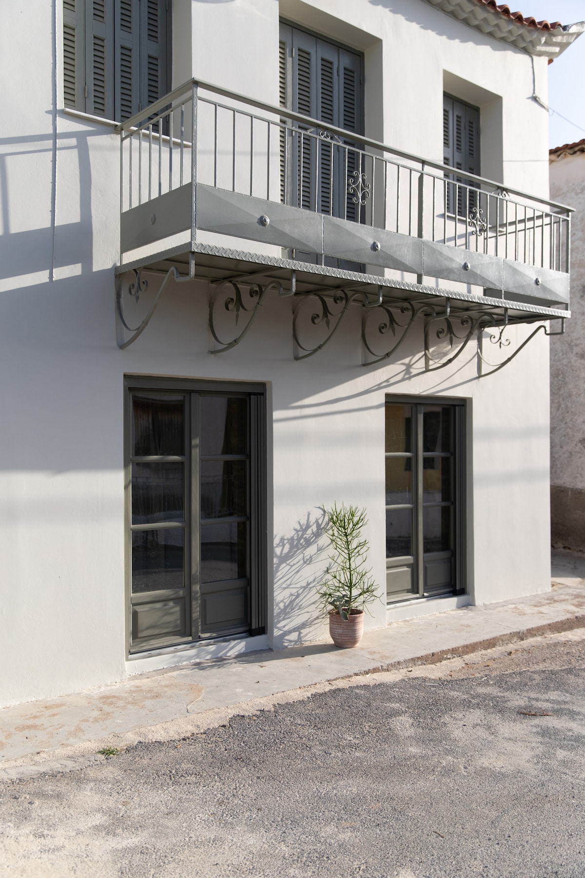 A two-story building with a neutral-toned exterior features a small balcony adorned with a decorative railing. Two large windows on the ground floor are framed in dark tones, while a potted plant adds a touch of greenery to the entrance.