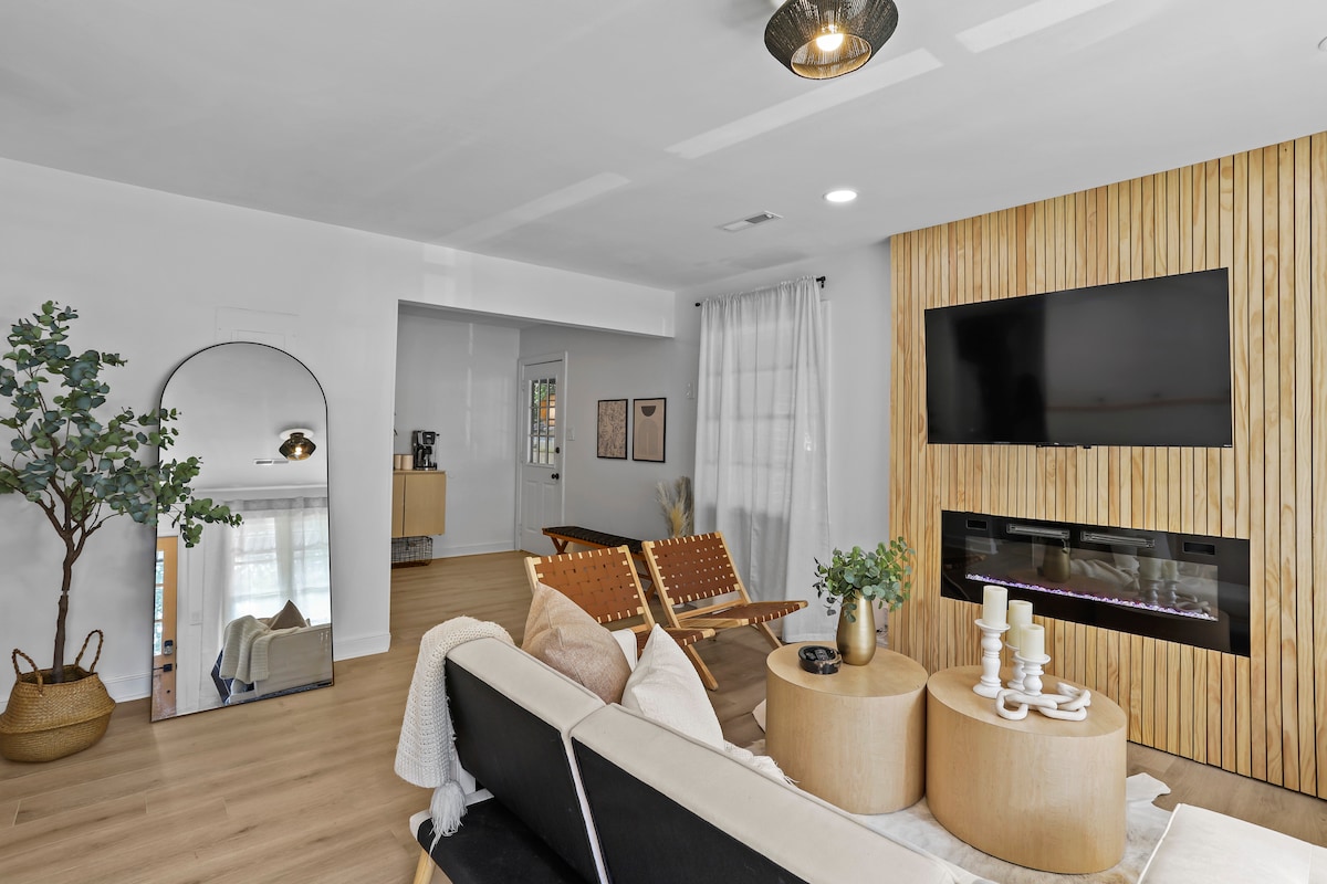 A stylish living area features a comfortable sofa positioned in front of a modern fireplace and wall-mounted TV. Natural light enters through large windows, illuminating wooden accents and contemporary furnishings. A decorative mirror and potted plant add subtle touches to the inviting space.