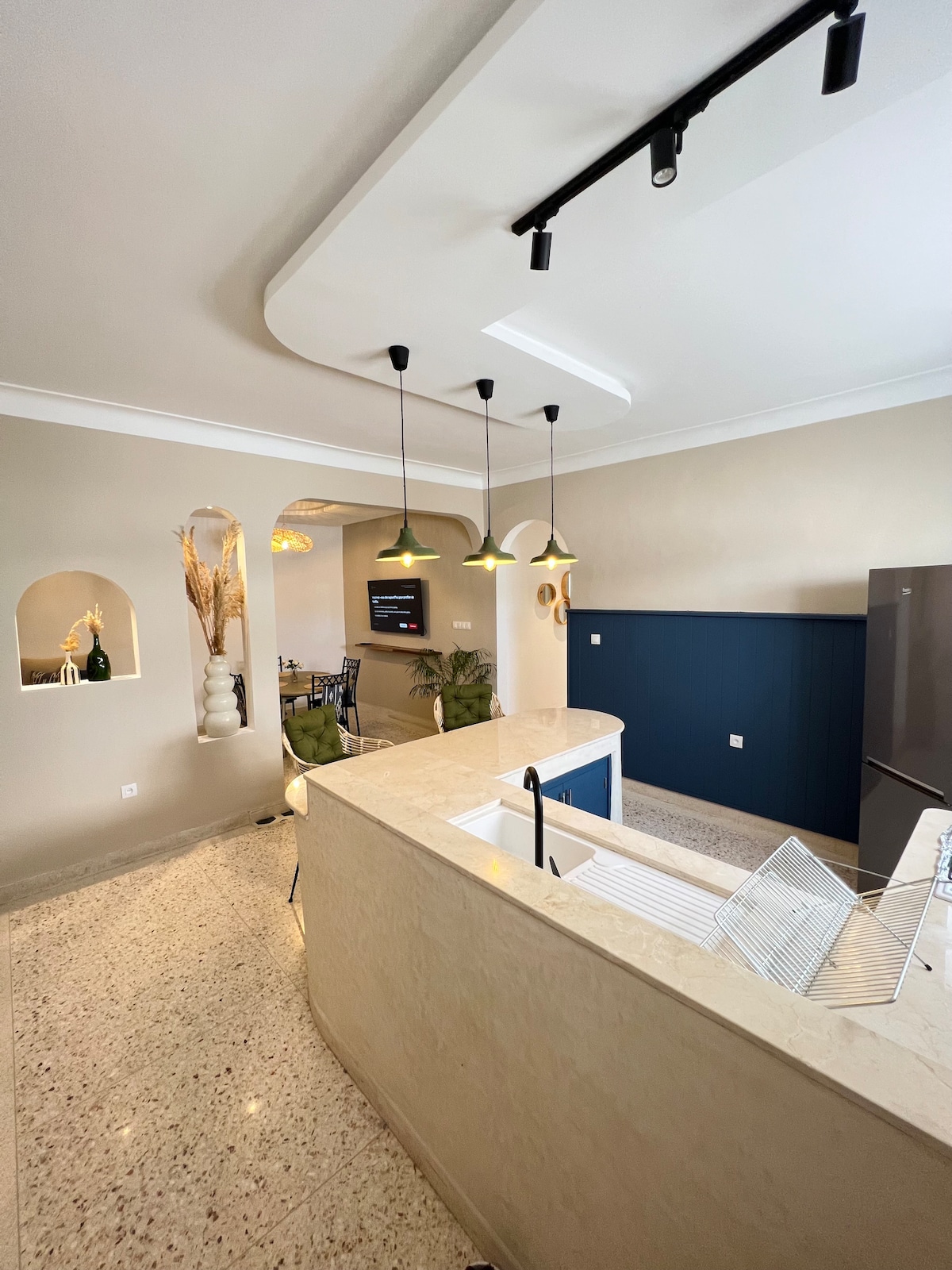 A modern kitchen featuring a spacious countertop with an integrated sink and a dish drying rack. Minimalist pendant lights are suspended above, while textured walls blend light and dark tones. An archway leads to a dining area, enhancing the open layout.