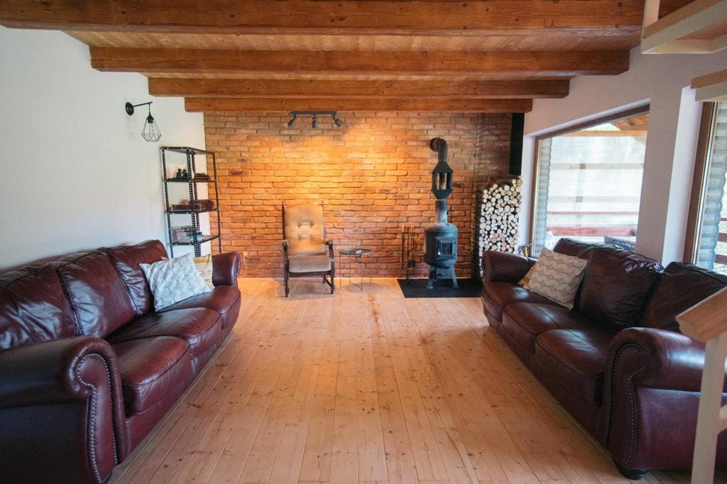 A spacious living area features two leather sofas arranged to create a cozy gathering space. A rustic brick wall serves as a backdrop to a small wood-burning stove, while a wooden chair and a shelving unit add character to the room.