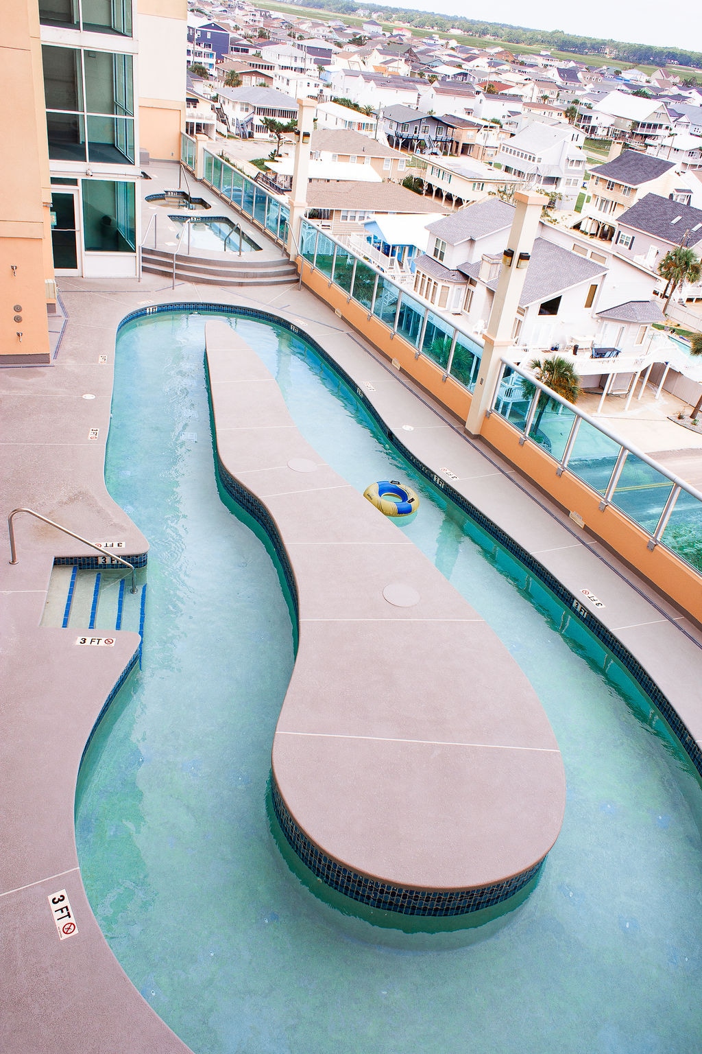 A winding lazy river pool is featured on the property, surrounded by a spacious deck. Lounge chairs are positioned along the sides, with an expansive view of the neighborhood rooftops in the background. The pool water appears calm and inviting.