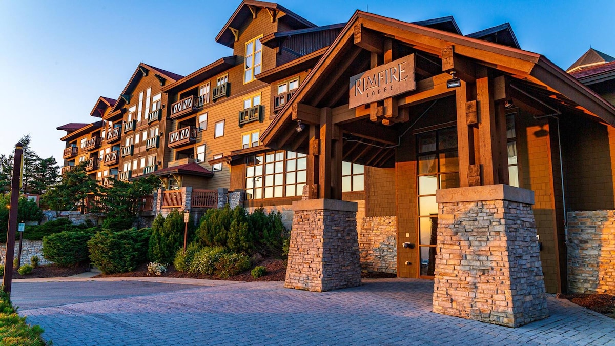 The exterior of Rimfire Lodge is presented with a blend of rustic wood and stone elements. The entrance features a grand overhang supported by sturdy pillars, providing a welcoming aesthetic. Lush landscaping surrounds the building, complemented by a blue sky and soft lighting during sunset.