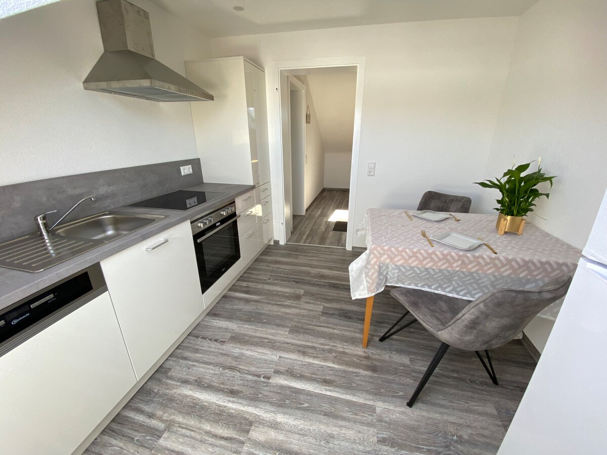 The kitchen area features sleek white cabinetry and modern appliances, including an oven and stovetop. A small dining table is set for two, accompanied by a potted plant. Natural light floods the space, highlighting the gray wood-style flooring.