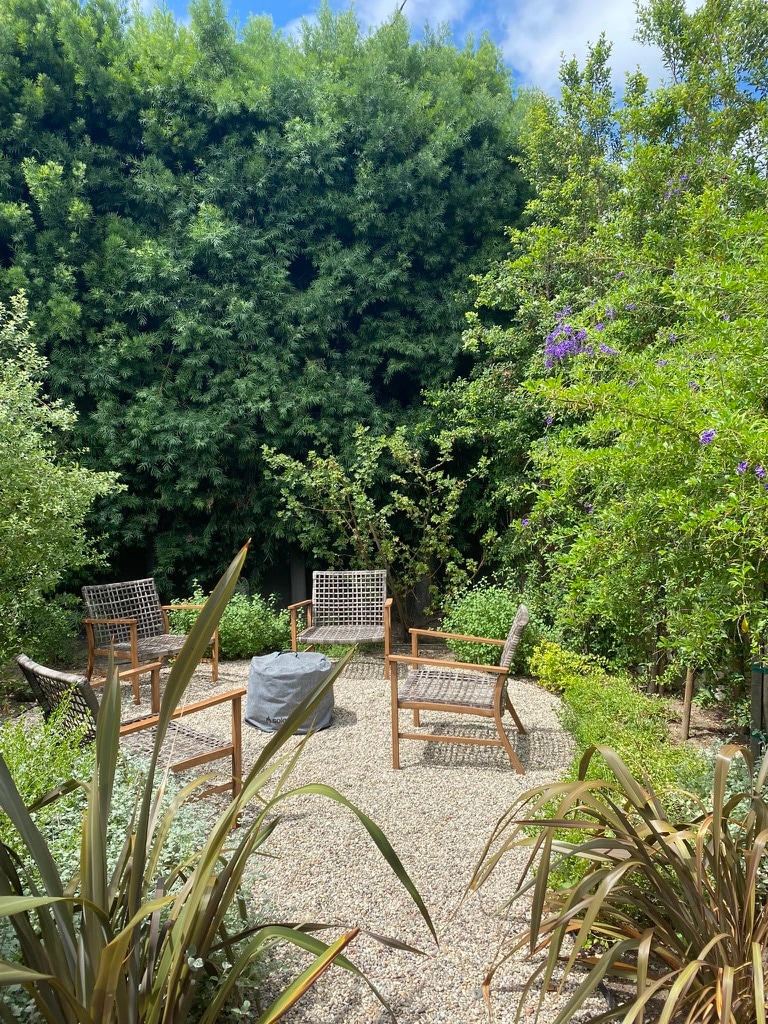 An outdoor seating area features four wooden chairs arranged around a central fire pit. Lush greenery surrounds the space, providing privacy and a serene atmosphere. The natural landscape includes various plants and bushes, creating a tranquil and inviting garden setting.