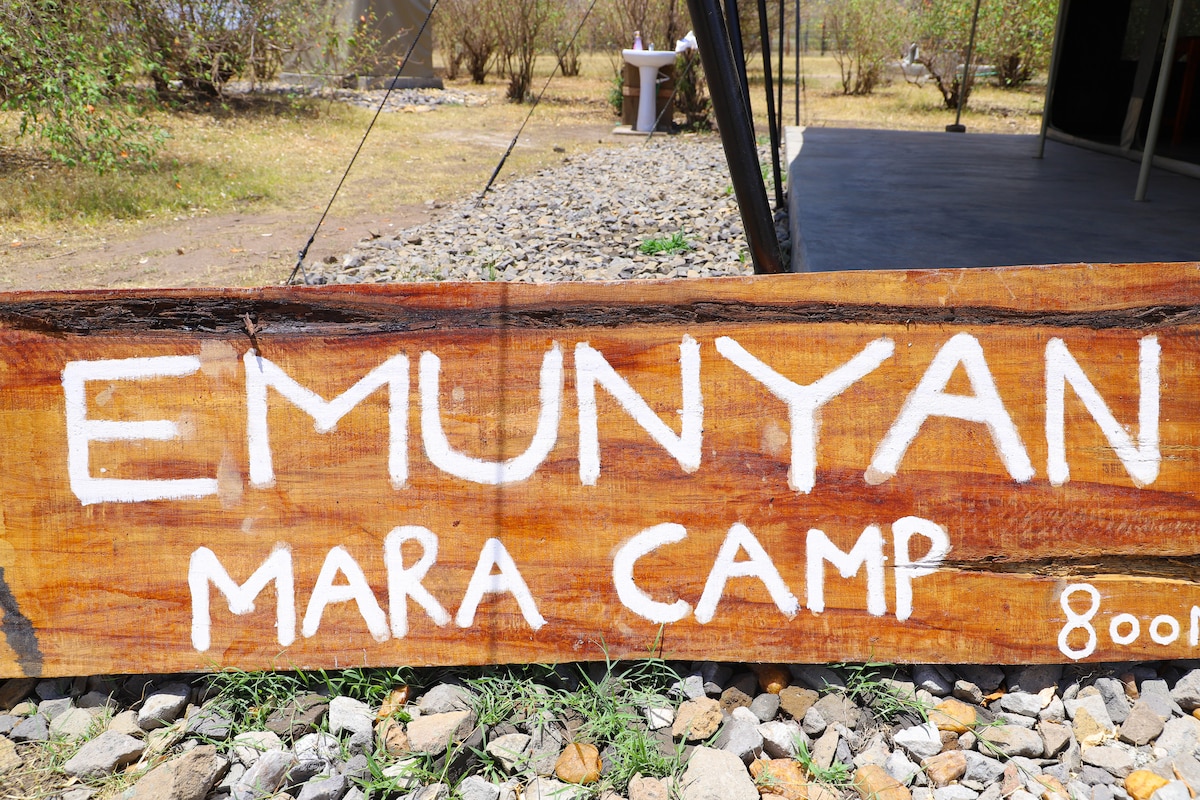A wooden sign displaying 'EMUNYAN MARA CAMP' is prominently placed on the ground, surrounded by pebbles and grass. The camp provides a rustic charm with a natural landscape visible in the background.