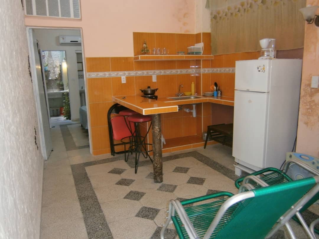 An open kitchen area features an L-shaped countertop with sink and cooking utensils. A compact refrigerator stands nearby. Two chairs provide seating, alongside a small table. Natural light enters through a door leading to an outdoor space, visible in the background.