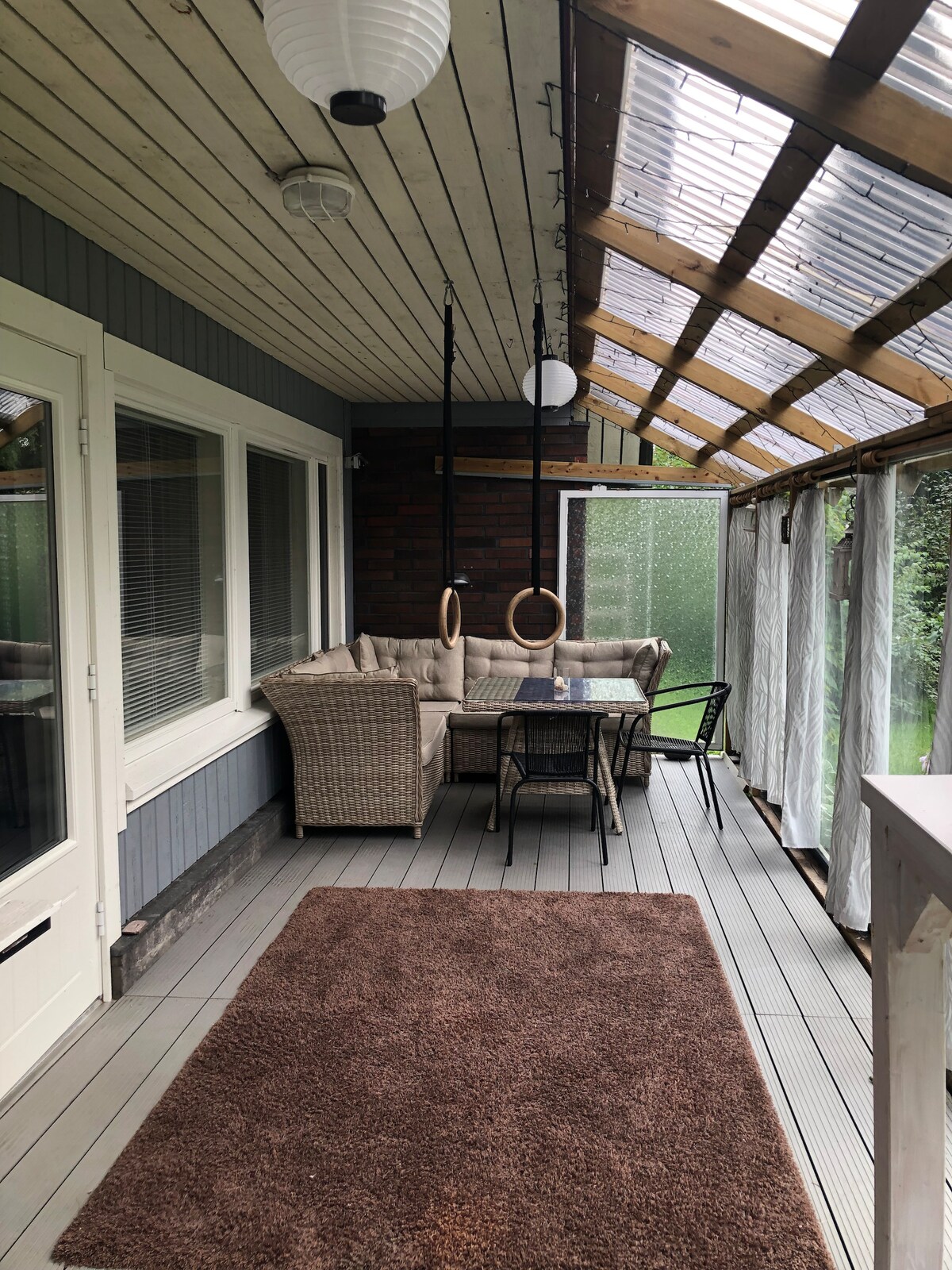A sunlit enclosed porch features a comfortable seating area with a cozy sofa and two hanging rings. A small dining table with chairs is present, positioned on a light-colored deck. Natural light filters through the glass roof and partly sheer curtains.