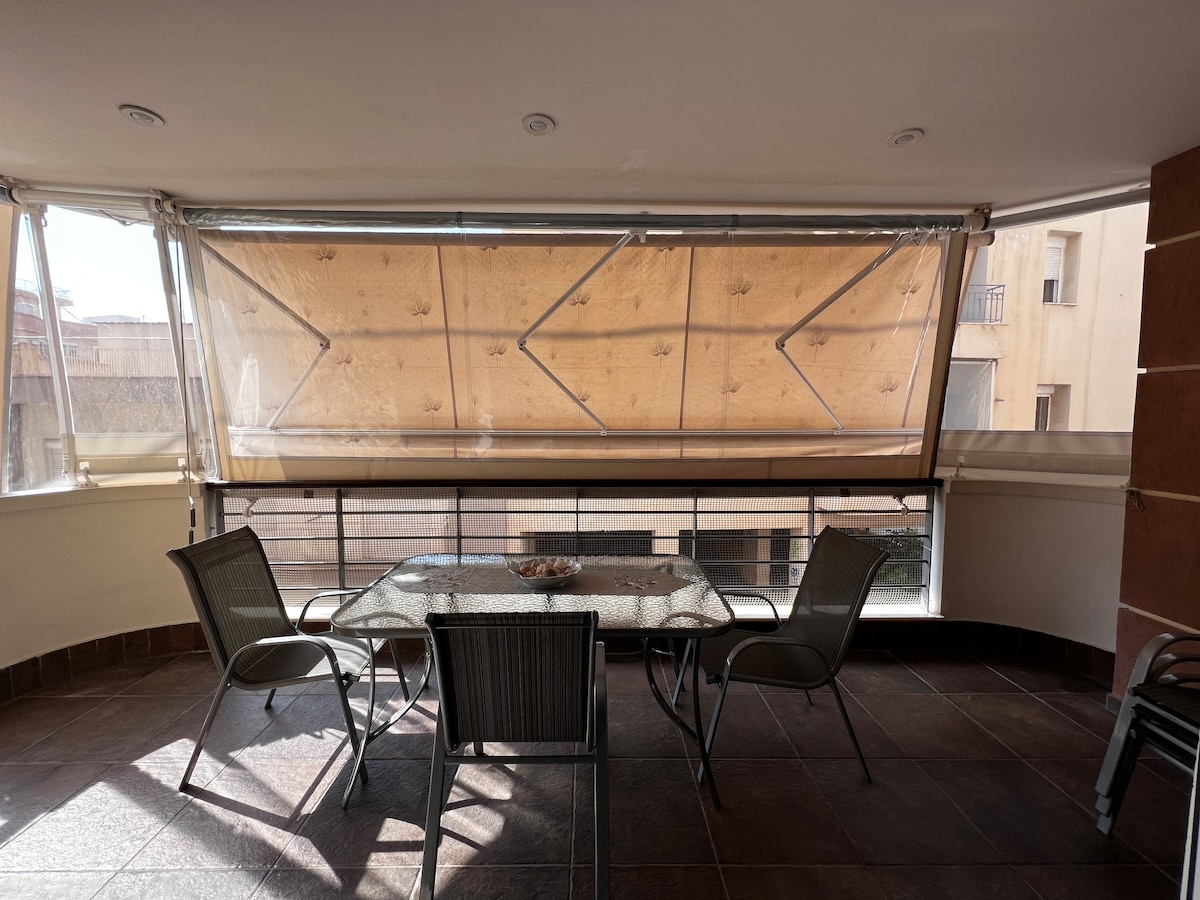 A covered outdoor terrace is shown, featuring a glass table surrounded by four chairs. Large windows allow natural light to enter, while shades are partially drawn, providing a sense of privacy. The surrounding walls are adorned with a warm tone, complementing the tiled floor.