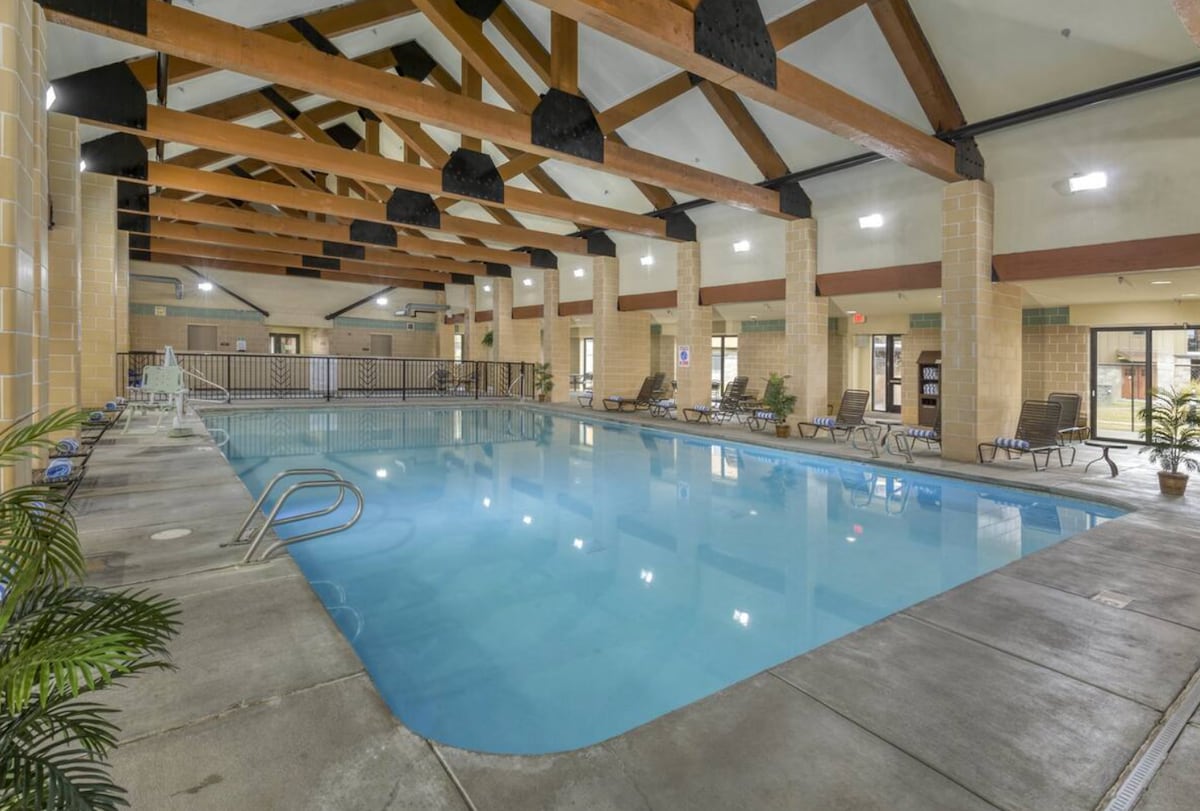 An indoor swimming pool is highlighted by exposed wooden beams overhead. Chairs are arranged around the pool deck, and tropical plants are placed intermittently along the perimeter. Soft lighting illuminates the serene water, creating a calm atmosphere.