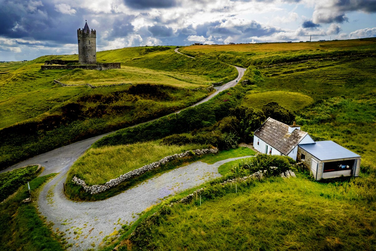 A scenic view captures the cottage nestled in lush green fields, with a winding gravel road leading to it. The 14th-century Doonagore Castle is visible in the background, surrounded by rolling hills under a dynamic sky filled with clouds.