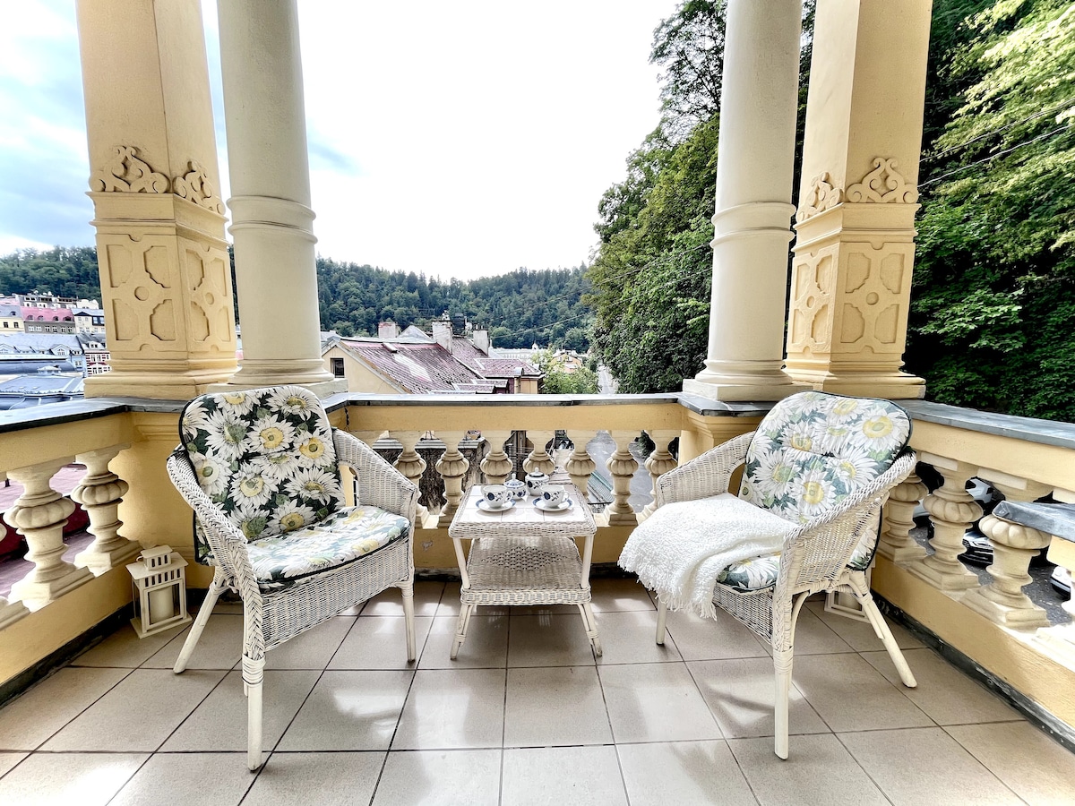 A balcony features two white wicker chairs adorned with floral cushions and a small table. A cozy blanket drapes over one chair. The view captures lush greenery framed by architectural columns, providing a serene outdoor space for relaxation.