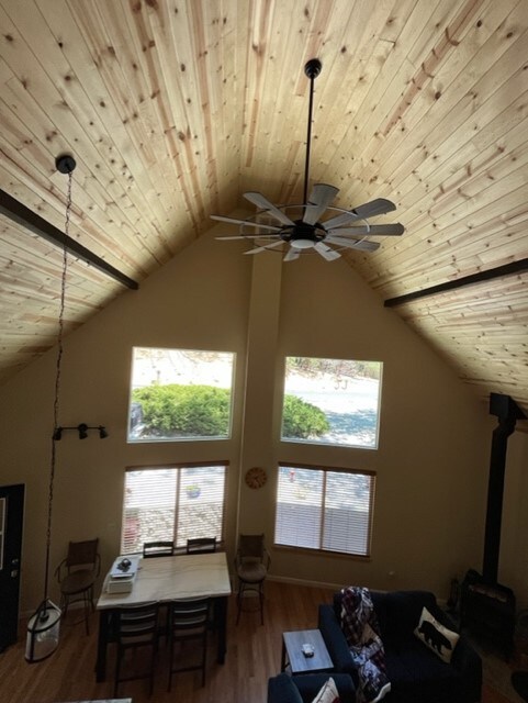An open living area showcases a high ceiling with wooden beams and a large ceiling fan. Bright windows provide natural light and views of the outdoor space. A wooden dining table is centrally located, surrounded by comfortable seating, creating a welcoming atmosphere.