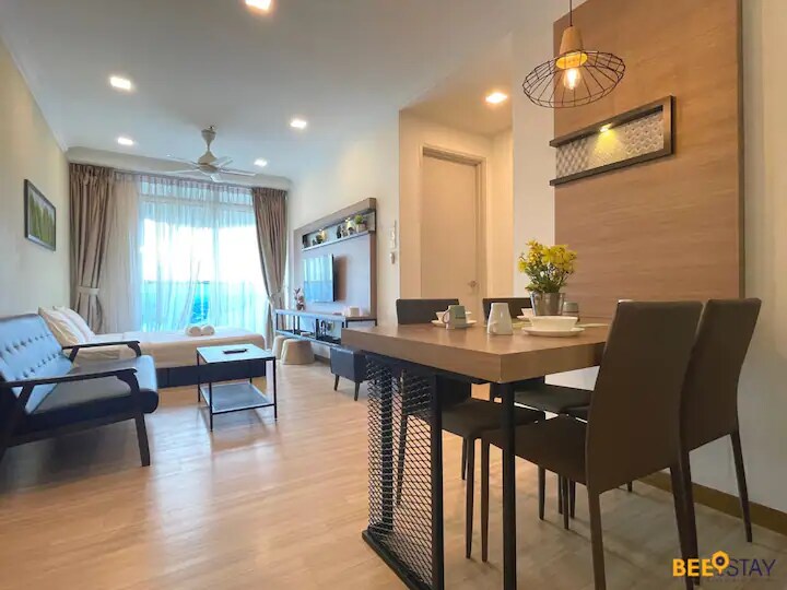 A spacious living area is highlighted by natural light streaming through large windows. A dining table with four chairs is visible, accompanied by decorative elements such as a small vase of flowers. A comfortable seating arrangement is positioned nearby, creating a welcoming space.