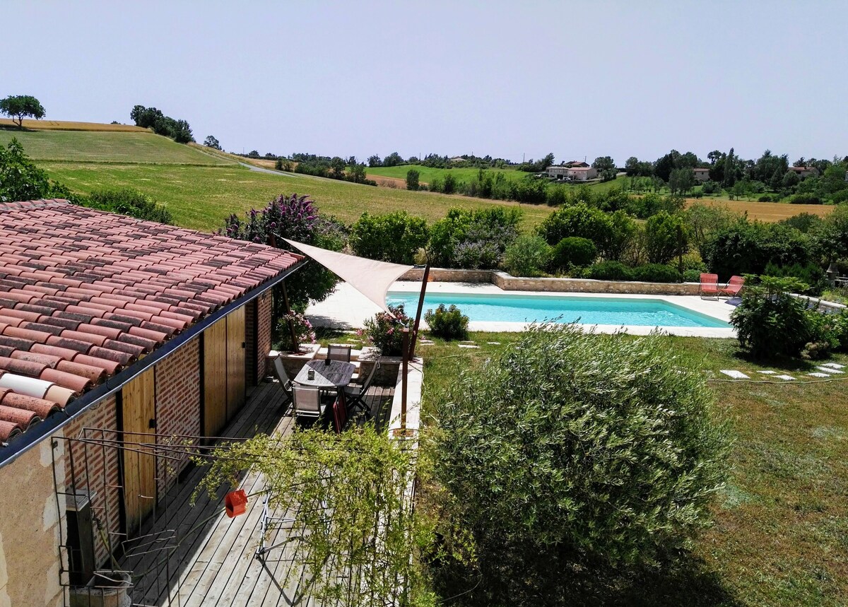 An outdoor area featuring a wooden terrace with a shade sail and seating arrangement. In the background, a shared salt-water pool is visible, surrounded by lush greenery and garden flowers. Rolling hills extend into the distance under a clear sky.