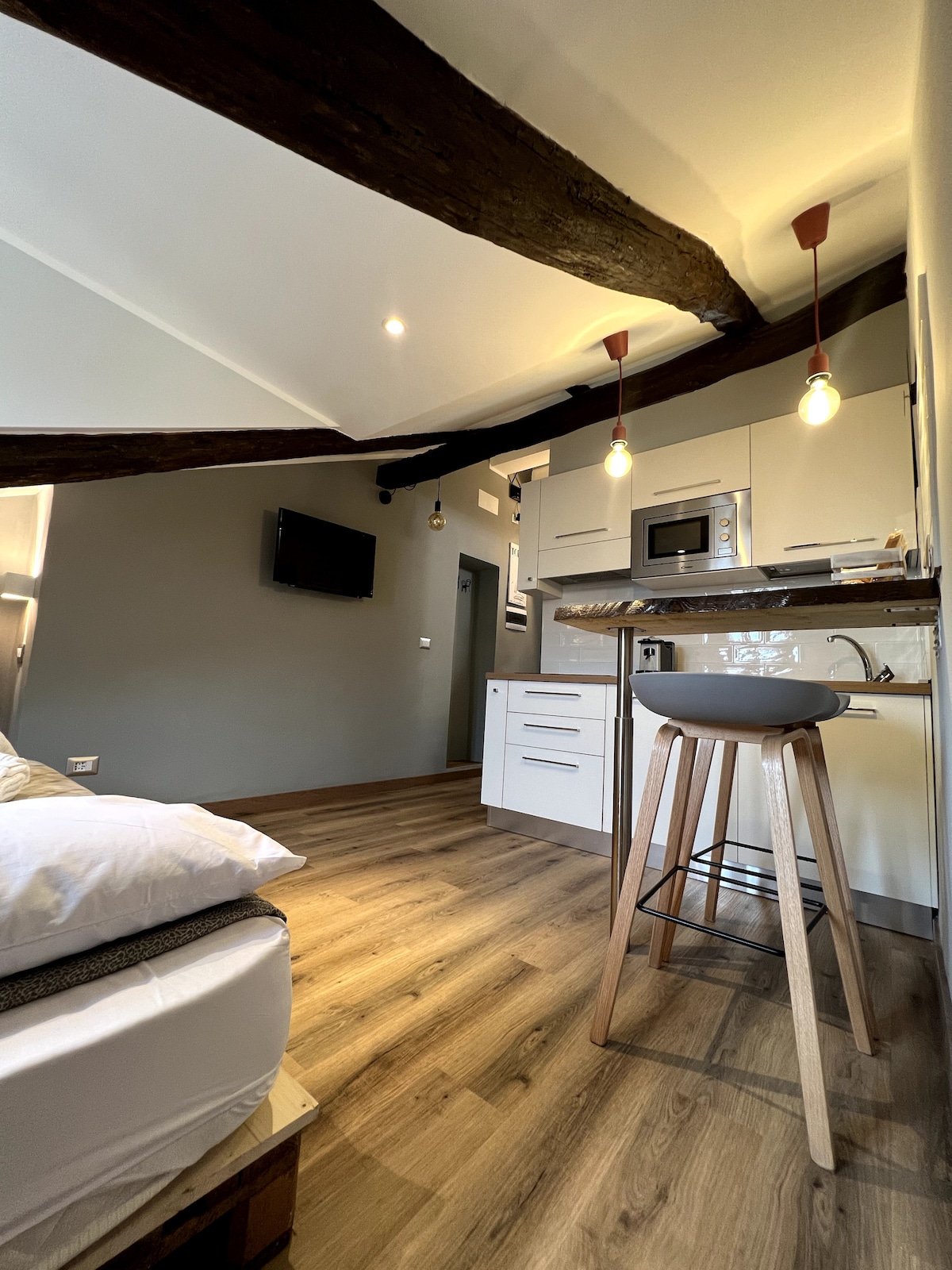 An open layout showcases a cozy sleeping area with a spacious bed in the foreground. A modern kitchenette features sleek cabinetry and bar stools. Exposed wooden beams adorn the ceiling, while soft lighting highlights the inviting atmosphere of the Loft.