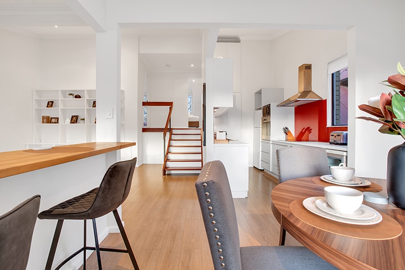 An open-plan kitchen and dining area is featured, showcasing a sleek modern design. A wooden breakfast bar with high stools separates the dining space, which includes a round table set with two cups. Light wood flooring enhances the bright atmosphere.