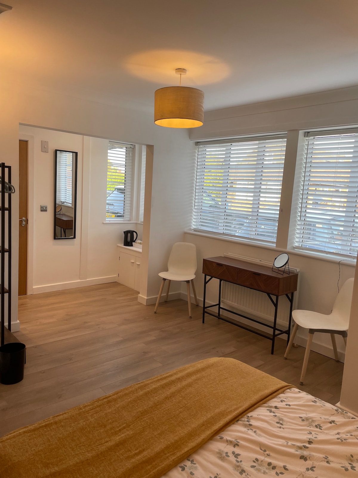 A bright and inviting room features large windows with white blinds, allowing natural light to fill the space. A wooden desk with a mirror sits beside two white chairs. A kettle on a countertop adds convenience, while light-colored flooring enhances the room's airy feel.