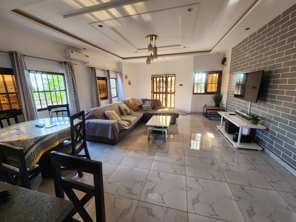 A spacious living area is showcased with comfortable seating arrangements, including a sectional sofa and a coffee table. Natural light is filtered through multiple windows, creating a bright ambiance. A dining table is situated near the kitchen area, and a television is mounted on the feature wall.