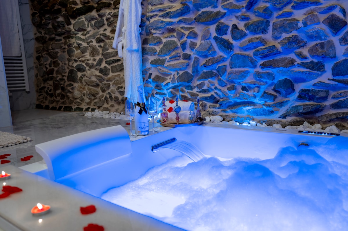 A spacious whirlpool is positioned against a stone wall, softly illuminated with blue LED lights. Bubbles fill the tub, surrounded by decorative rose petals. A plush white robe hangs nearby, and two bottles sit on the edge, enhancing the serene ambiance.