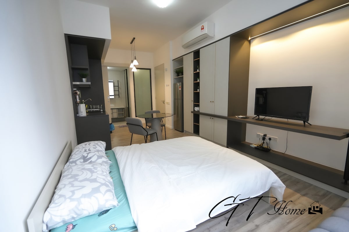 A modern studio unit is presented, featuring a spacious bed with a light-colored blanket. A functional kitchenette is visible on one side, while a Smart TV is mounted on the wall opposite. Natural light enters through a nearby window, creating a bright atmosphere.