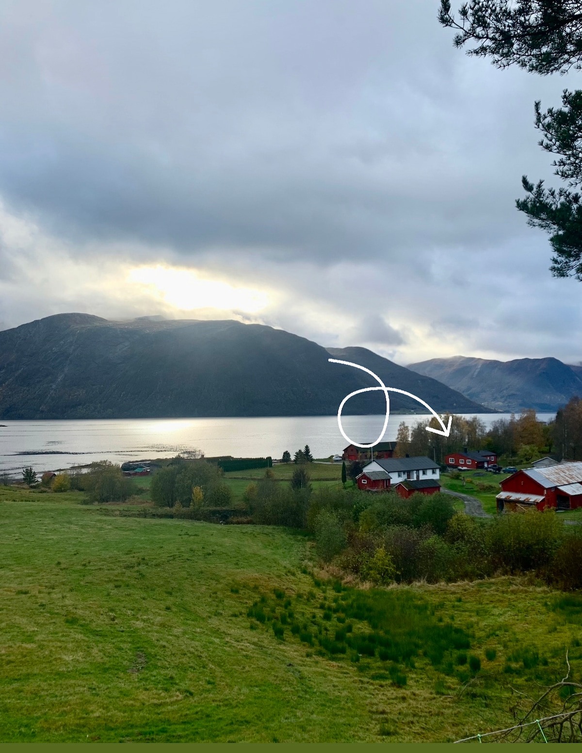 A scenic view captures a peaceful landscape featuring mountains and a serene lake. Various structures, including a red house and outbuildings, are visible in the foreground, surrounded by greenery. Soft clouds diffuse the sunlight, creating a tranquil ambiance in the setting.