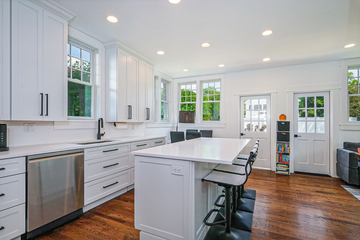 A modern kitchen is displayed with white cabinetry and stainless steel appliances. A large central island with seating faces a row of windows. Warm wooden flooring complements the bright, inviting space, which features sleek fixtures and contemporary design elements.