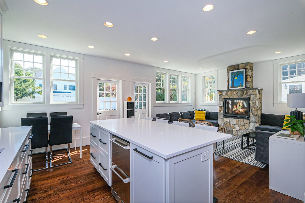 A bright and airy kitchen space features white cabinetry and a large island topped with sleek countertops. Surrounding windows allow natural light to illuminate the room. A cozy seating area with a stone fireplace is visible, and modern furnishings complement the overall design.