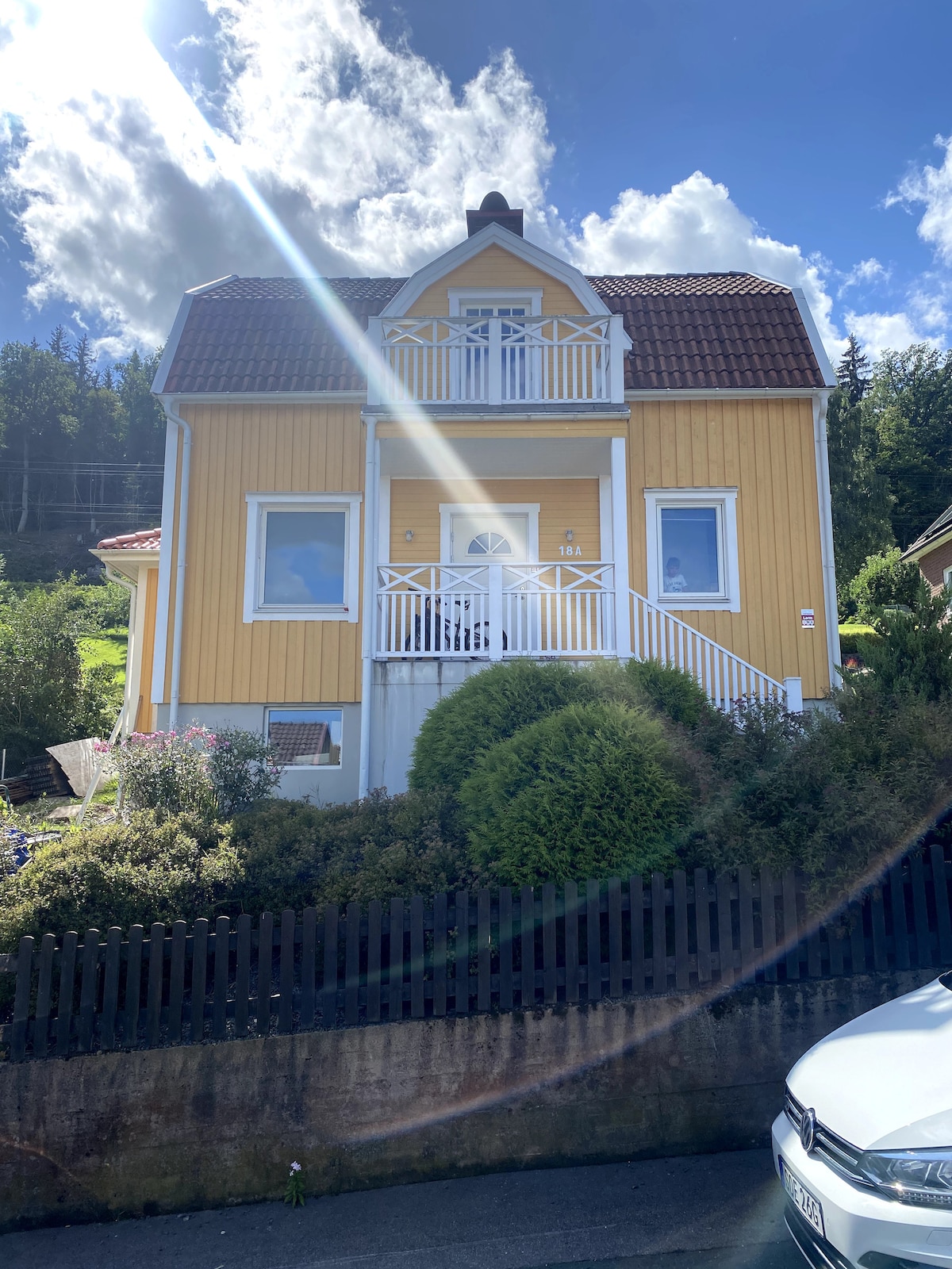 A charming yellow house is visible, featuring a traditional design with a gabled roof and a front balcony. Lush greenery surrounds the property, complemented by a neatly trimmed hedge and garden. The clear blue sky and scattered clouds enhance the inviting exterior.