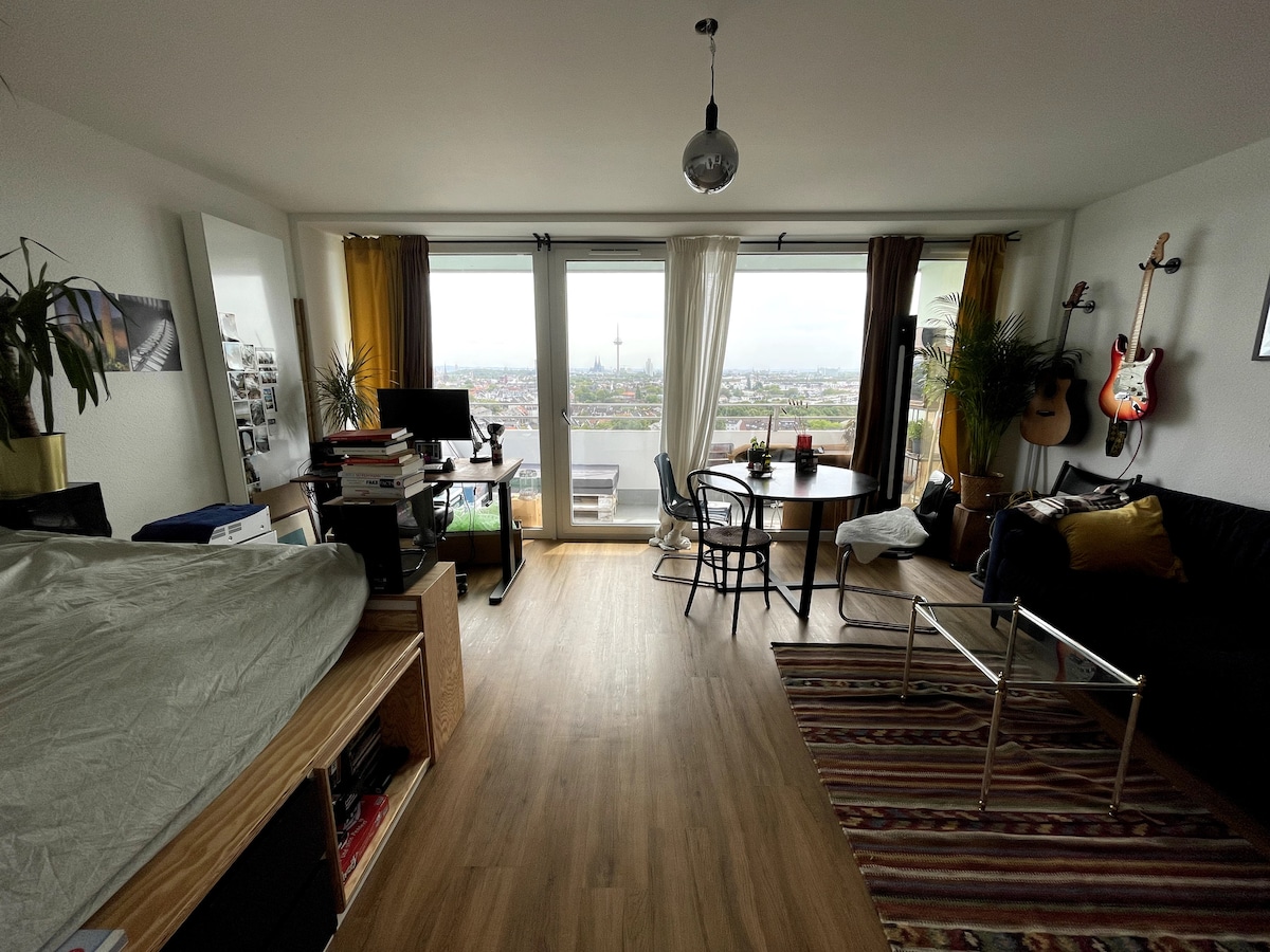 A bright living space is presented, featuring a cozy sleeping area with a bed and a black sofa. A round table surrounded by chairs and a work desk can be seen. Floor-to-ceiling windows offer an expansive view of the city, while decorative plants add a touch of nature.