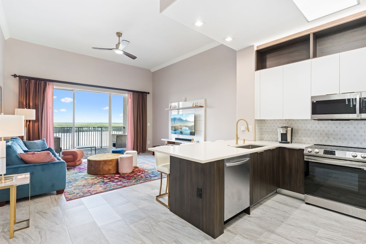 A modern suite features an open layout with a well-equipped kitchen and living area. Large glass doors provide access to a balcony with a scenic view. A colorful rug enhances the space, while sunlight fills the room, highlighting the contemporary furnishings.