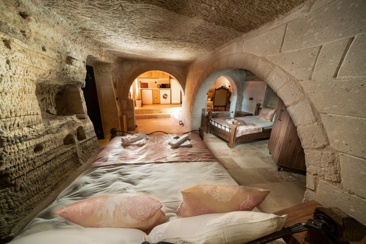 A spacious cave suite features natural stone walls with arched openings leading to distinct sleeping areas. Soft bedding and decorative pillows complement the neutral tones. A wooden dressing table is positioned near the entrance, with warm lighting accentuating the unique cave architecture.