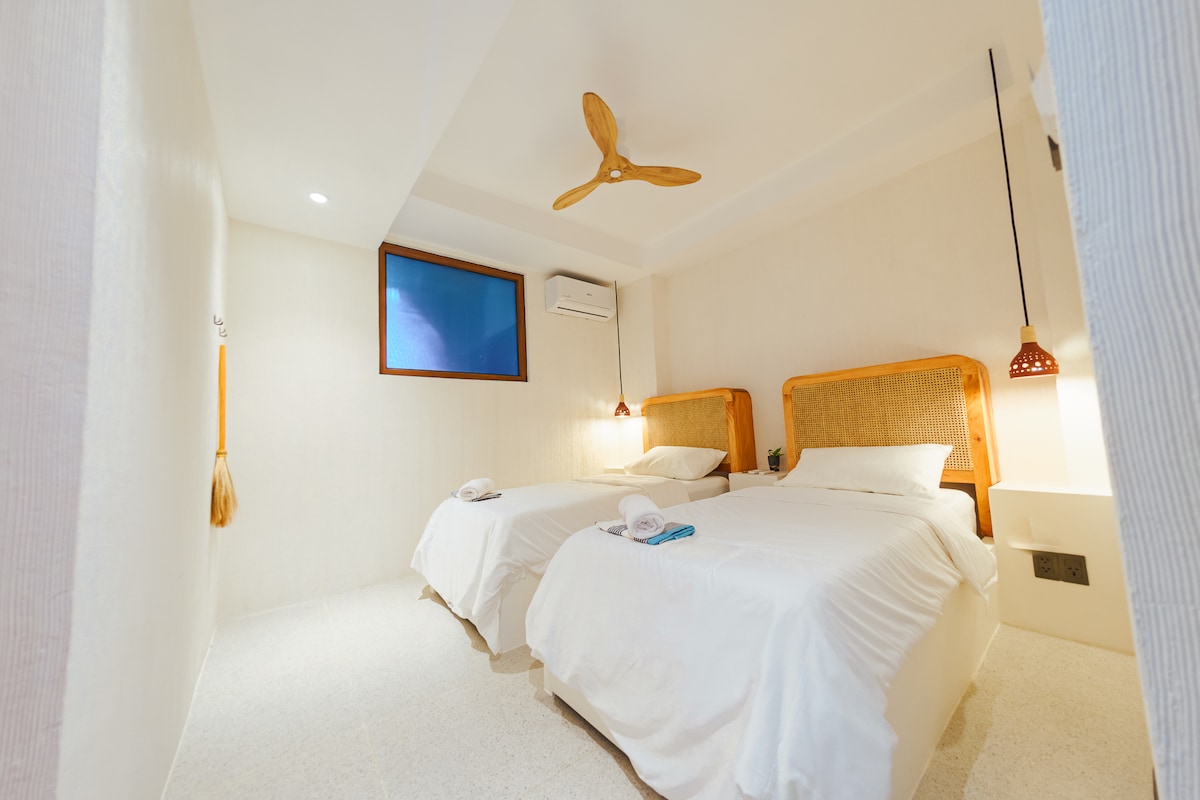 A minimalist room is furnished with two spacious beds, complemented by light bedding. Natural light enters through a window, while a ceiling fan adds comfort. Decorative light fixtures hang above each bed, enhancing the room's functional design.