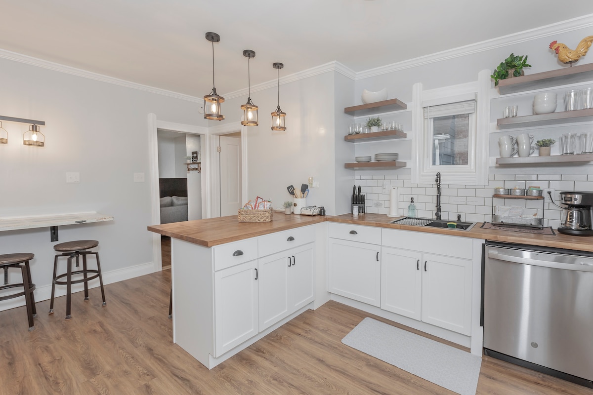 A modern kitchen is seen, featuring white cabinetry and a wooden island with seating for two. Pendant lights illuminate the space, which includes stainless steel appliances, open shelving with decor, and a black sink. Light-colored walls complement the warm wood flooring.