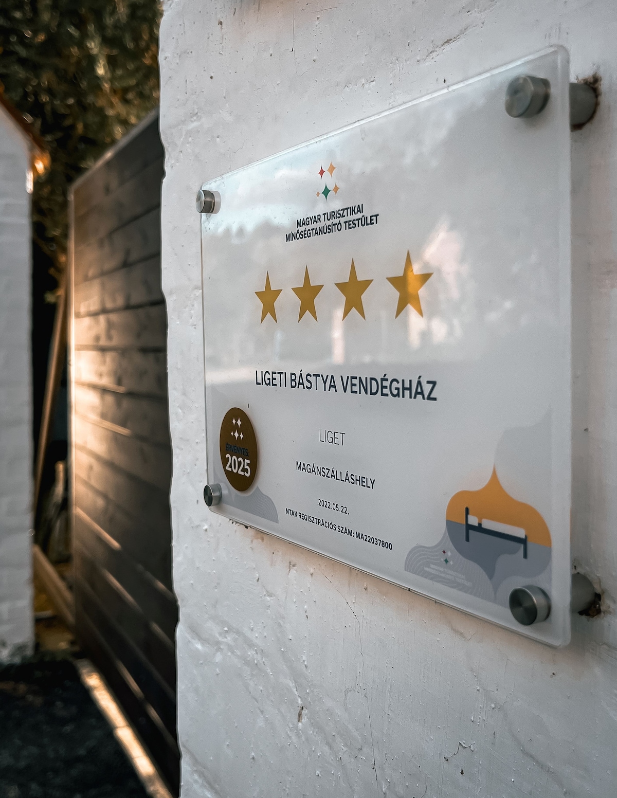 A plaque is displayed on a white wall, featuring four gold stars and the name 'Ligeti Bástya Vendégház.' The year 2025 is noted, indicating an award or certification. The surrounding area suggests a well-maintained rustic environment with natural elements.
