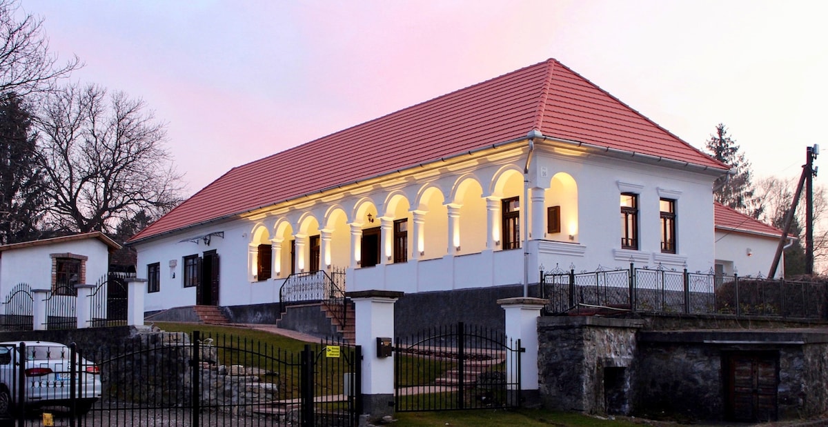 The exterior of Antik Vendégház is depicted, showcasing its traditional architectural style. The building features a red-tiled roof and multiple arched entries, with soft lighting highlighting its facade. A well-maintained garden and fenced area are visible in the foreground.