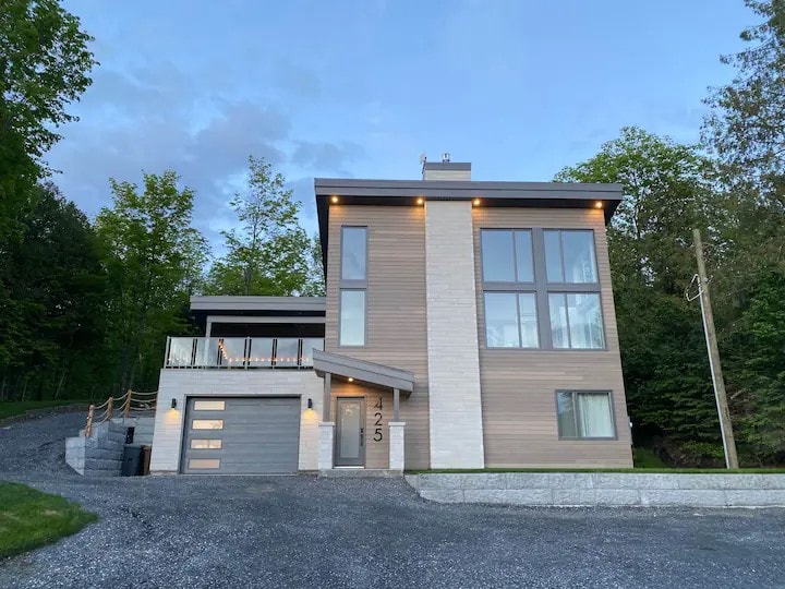 The modern chalet is set in a natural landscape, featuring a blend of wood and stone. Large windows allow for ample natural light, and a balcony is visible above the entrance. A gravel driveway leads to a garage with a minimalist design.