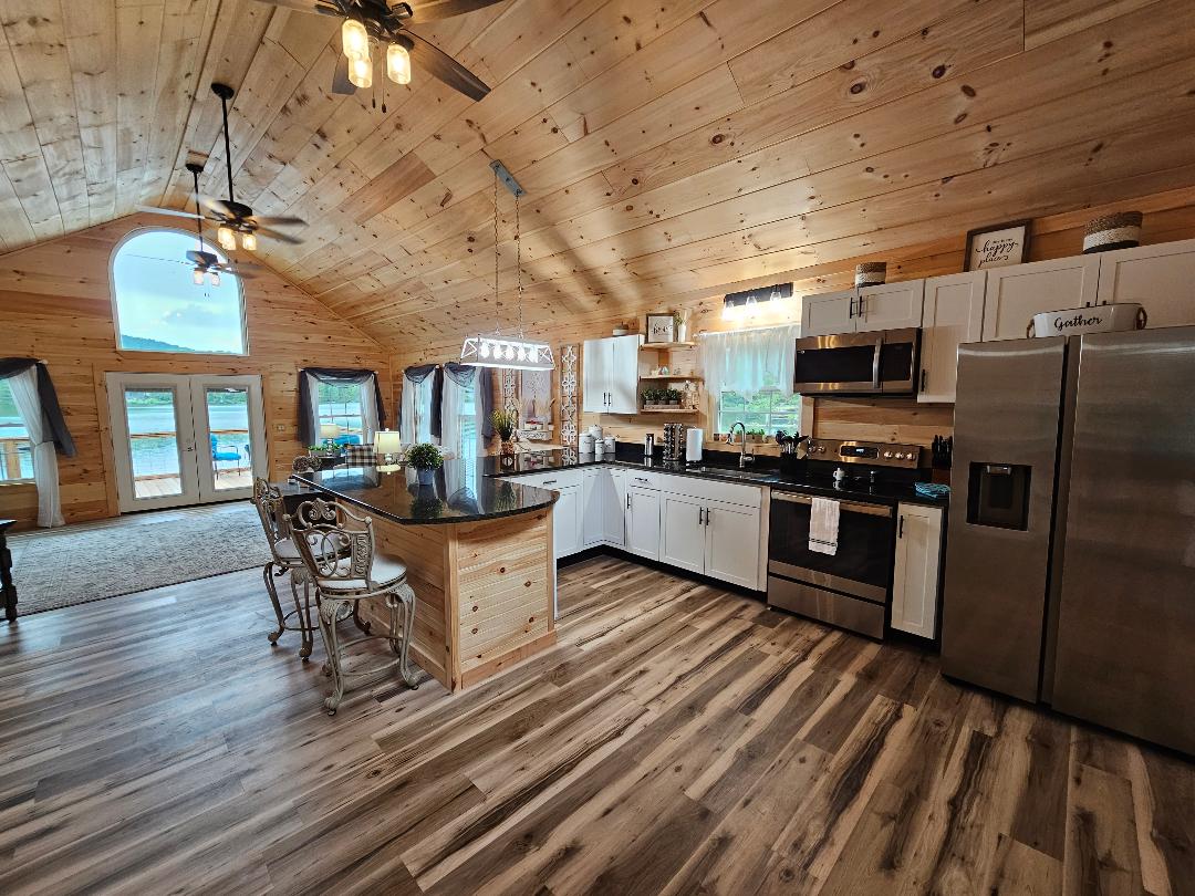 A spacious kitchen is highlighted with light wood paneling and modern appliances. A large island provides seating for four. Windows bring in natural light, showcasing views of the water. Nearby, a dining area features a decorative chandelier and additional seating.