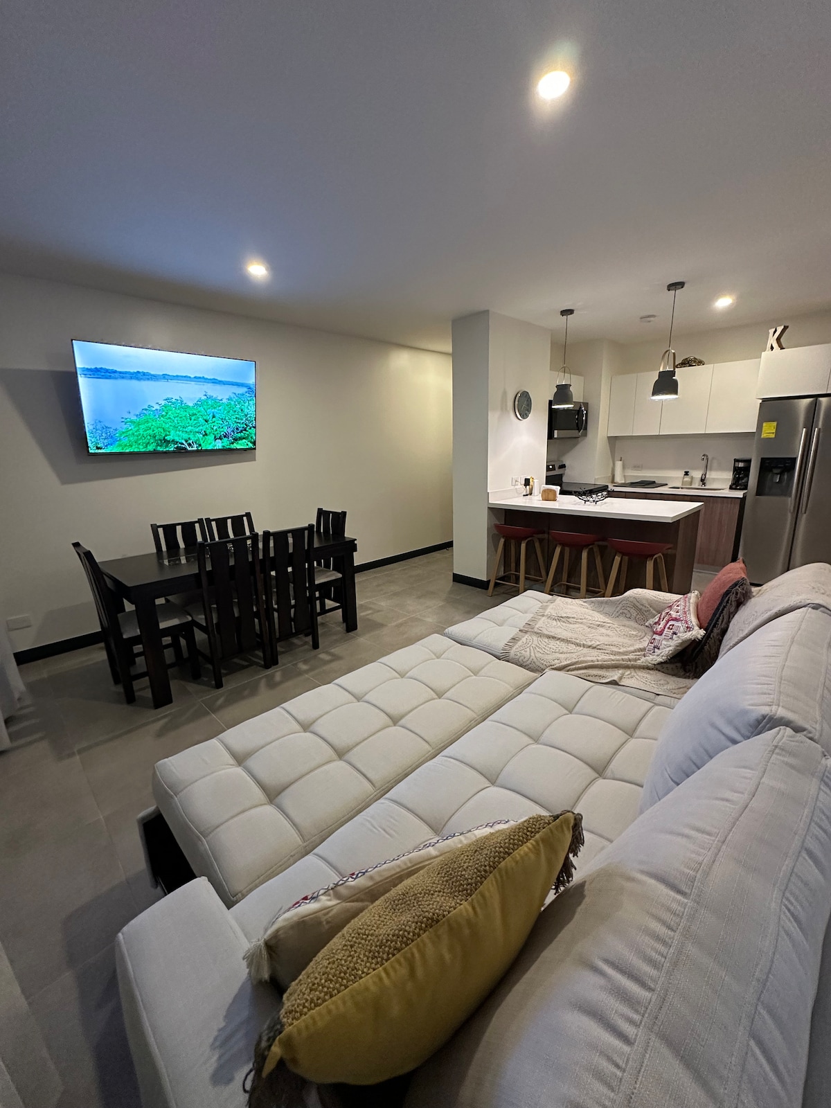 A modern living area is showcased, featuring a comfortable sofa with decorative pillows. A dining table with six chairs is positioned nearby, and a wall-mounted television is visible. The kitchen area includes sleek cabinetry and appliances, creating a cohesive and functional space.