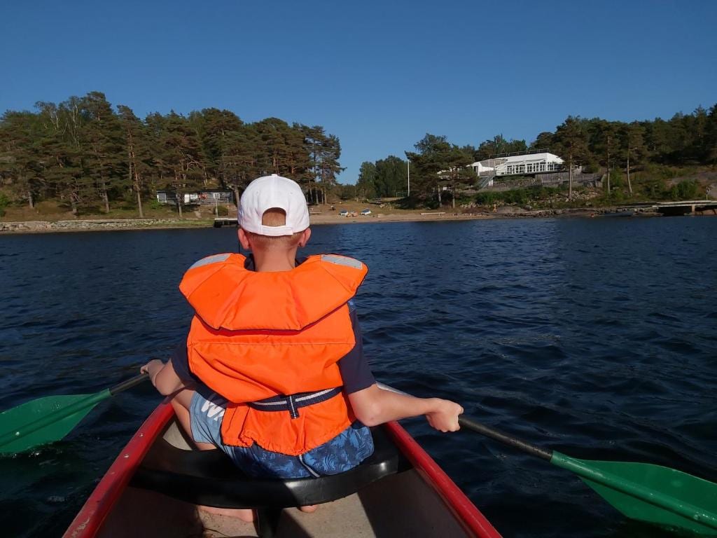 A child is seated in a canoe, wearing an orange life jacket, as they paddle through calm waters. Dense trees line the shore in the background, with a glimpse of a white building visible. Bright sunlight reflects off the water's surface.