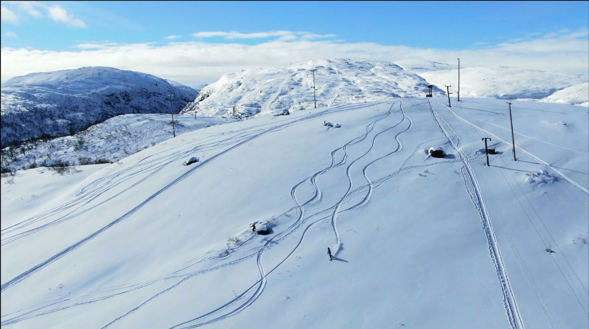 A snowy landscape is captured, featuring a vast expanse of white terrain marked by ski tracks. Several hills are visible in the background, with ski lifts positioned along the slopes, enhancing accessibility for winter sports enthusiasts.