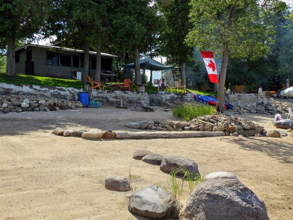 A sandy beach area is showcased in the foreground, with a rocky path leading towards the cabin. In the background, the cabin is surrounded by trees and outdoor seating, while a Canadian flag adds color to the serene setting.