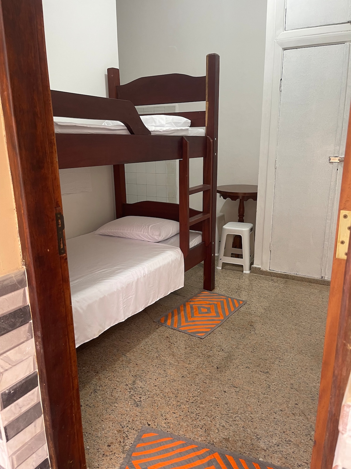 A simple sleeping area is presented, featuring a wooden bunk bed accompanied by a twin bed. The space displays a neutral color scheme with beige walls and a tiled floor. A small table and a white stool are positioned nearby, contributing to the functional layout.