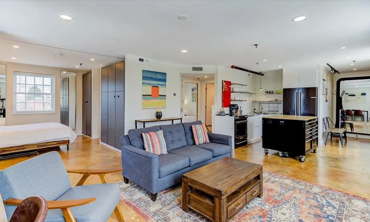 The spacious loft features an open layout with polished hardwood floors and bright decor. A comfortable sofa is positioned in front of a wooden coffee table, while a modern kitchen area with stainless steel appliances and storage cabinets is visible in the background.