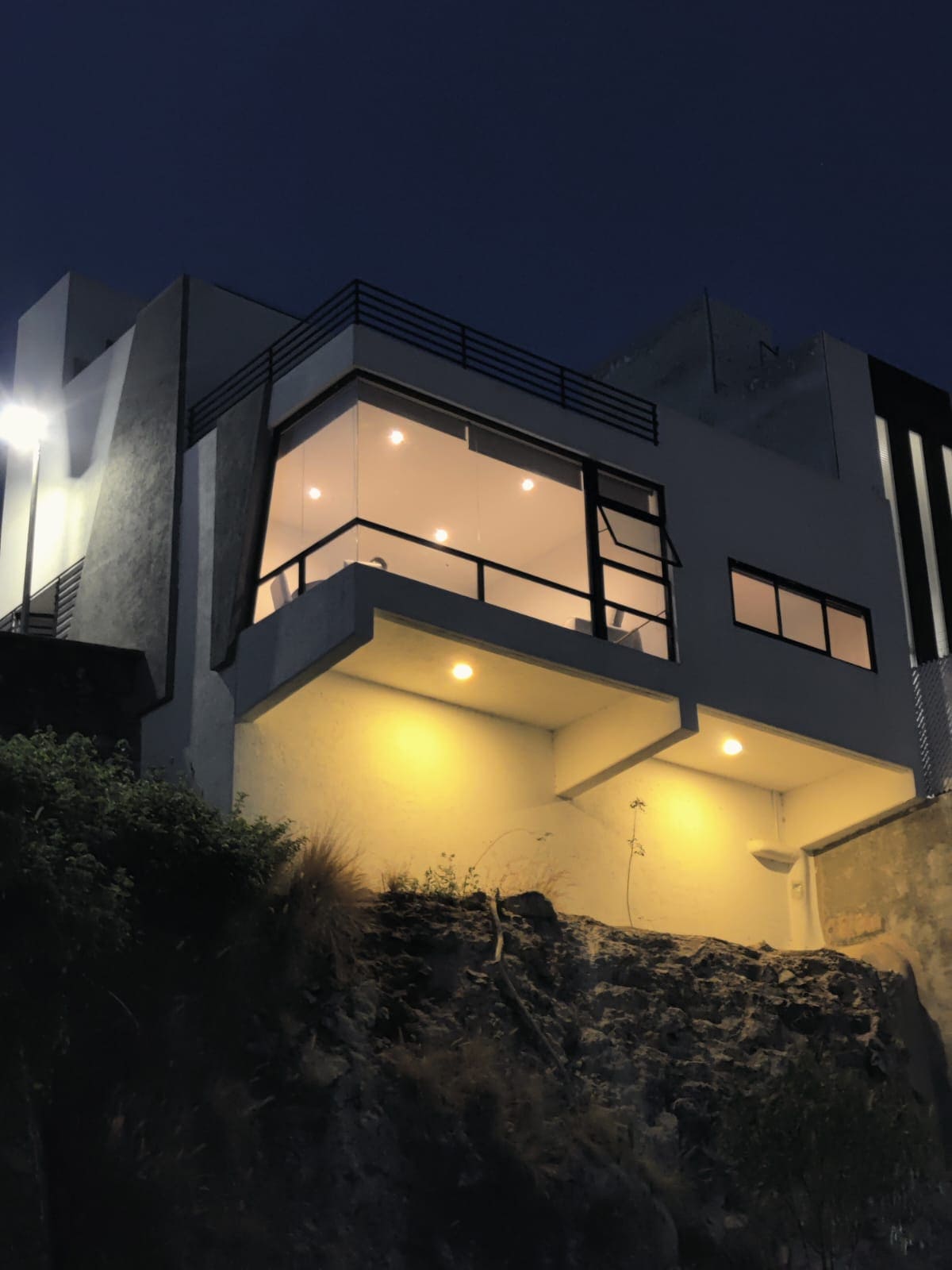 The exterior of the modern residence is highlighted by soft outdoor lighting, illuminating the architectural features at dusk. Large glass windows create a connection to the surrounding landscape, while the structure is set against a rocky terrain, providing a serene and private setting.